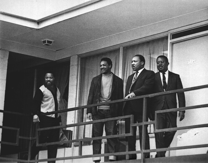 Four Black men stand in front of a railing in front of windows. The photo is in black and white. Two men on the right are in suits. The others are dressed more casually.