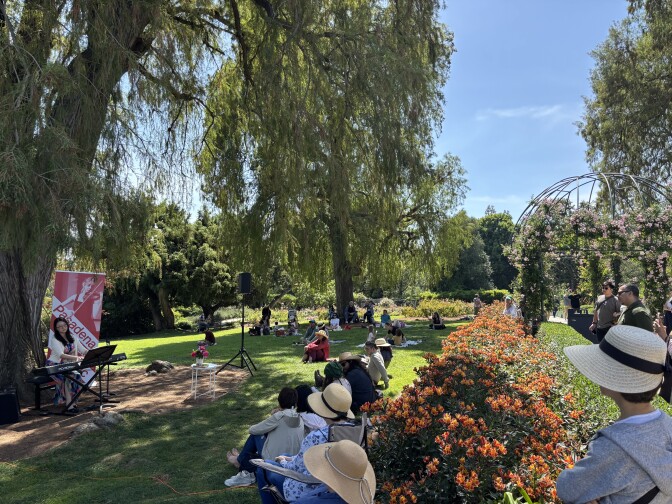 A scene of a garden with orange flowers and grass and large trees and a pianist playing an electronic keyboard under the tree. Onlookers sit on the grass and stand behind the orange flowers, many wearing straw sun hats.