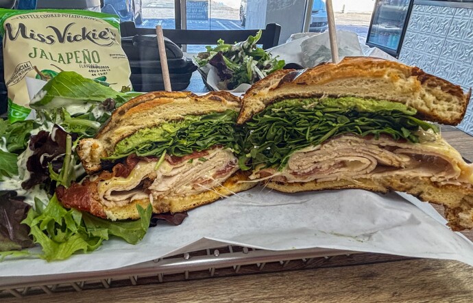 A large sandwich cut in half on a tray, filled with layers of sliced turkey, crispy bacon, leafy greens, melted cheese, and avocado spread on toasted bread. Served with a side salad and a bag of Miss Vickie's Jalapeño kettle-cooked chips.