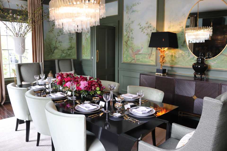 A formal dining room with a large glass chandelier hanging over a table that seats eight people. The table is set with plates, utensils and glasses, and has a pink and red floral centerpiece. There are painted murals on the walls and a large window behind the table. 