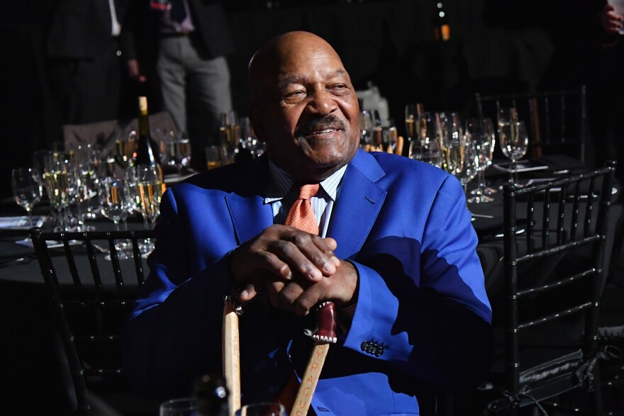 An older dark-skinned bald man with mustache, wearing a neon blue suit and salmon tie, is seated in front of a table set for a formal dinner.