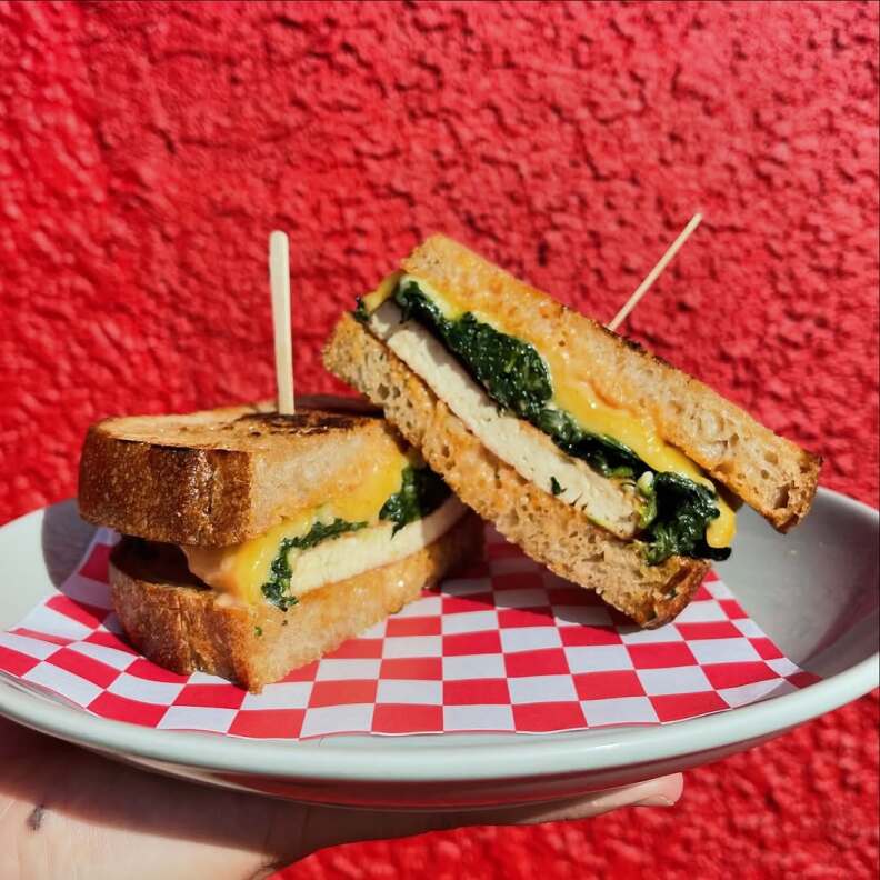 A tofu patty melt on a white plate, being held up in front of a red-painted wall. The cross-section of the sandwich is facing the camera. 
