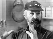 A bearded white man in a cap motions with his hands while looking off camera.