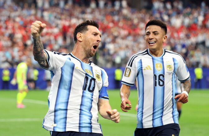 Two medium skinned men wearing blue and white soccer jerseys are smiling and punching the air on a soccer field.