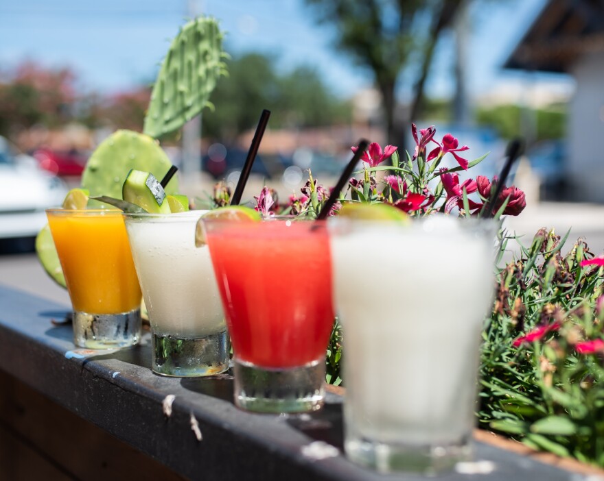 Four glasses filled with white, pink or orange liquid sit on a ledge, surrounded by some foliage.