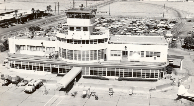 A black and white photo shows a white building flanked with bay windows and a tower on top. Cars are seen in the distance in a parking lot with the tale of an airplane seen in the lower left corner. 