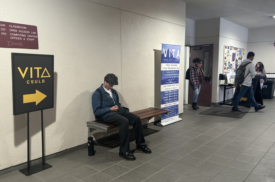 A handful of students with backpacks make their way in an out of a school building. In the foreground, a person with short dark hair sits on a bench, listening to music while scrolling through their cell phone. In the background, a bulletin board is teeming with flyers advertising campus resources and events. 