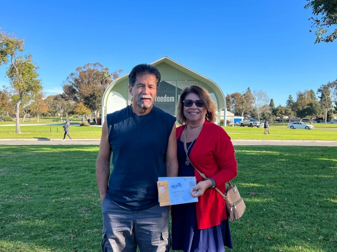 An older man with gray hair and goatee wearing a sleeveless navy blue shirt and gray shorts stands next to an older woman with short light brown hair, sun glasses, a navy dress, and red sweater. They stand on a patch of grass in a park. 
