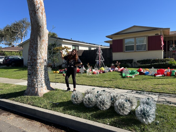 A woman wearing a black tshirt and leggings strings holiday lights around a tree. Other Christmas decorations are visible on her front yard. 
