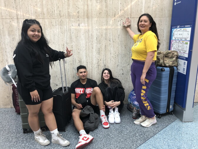 Four people with luggage. A young woman wearing a black sweatshirt and shorts smiles while standing at the left. On the right, her mother smiles and stands wearing a yellow Lakers shirt and purple Lakers sweatpants. At center a young man in a black t-shirt and young woman in black sweatshirt and pants sit on the ground, leaning against a marble wall, smiling. 