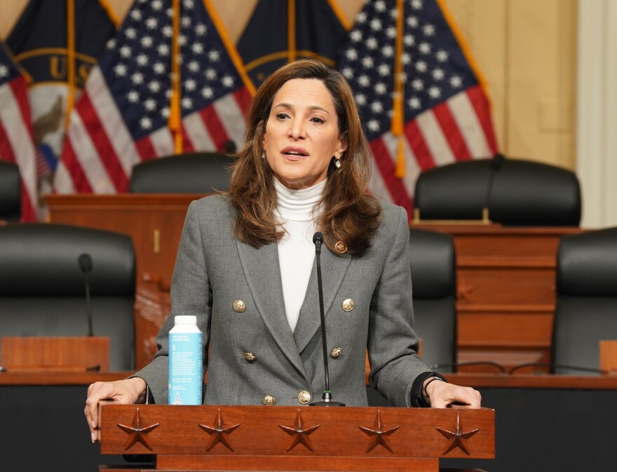 A woman wearing a grey suit soeaks into a microphone at a wooden podium. Behind here are two rows of empty, black leather seats and two American flags.