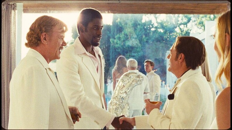 A group of people all in white outfits, men largely in suits, women in dresses. On the left is the actor portraying Dr. Jerry Buss with a drink in his hand. Next is the actor portraying Magic Johnson, who shakes hands with the actor portraying Donald Sterling. A blonde woman is seen from behind on the far right, with other people on the other side of glass in the center around a pool outside.