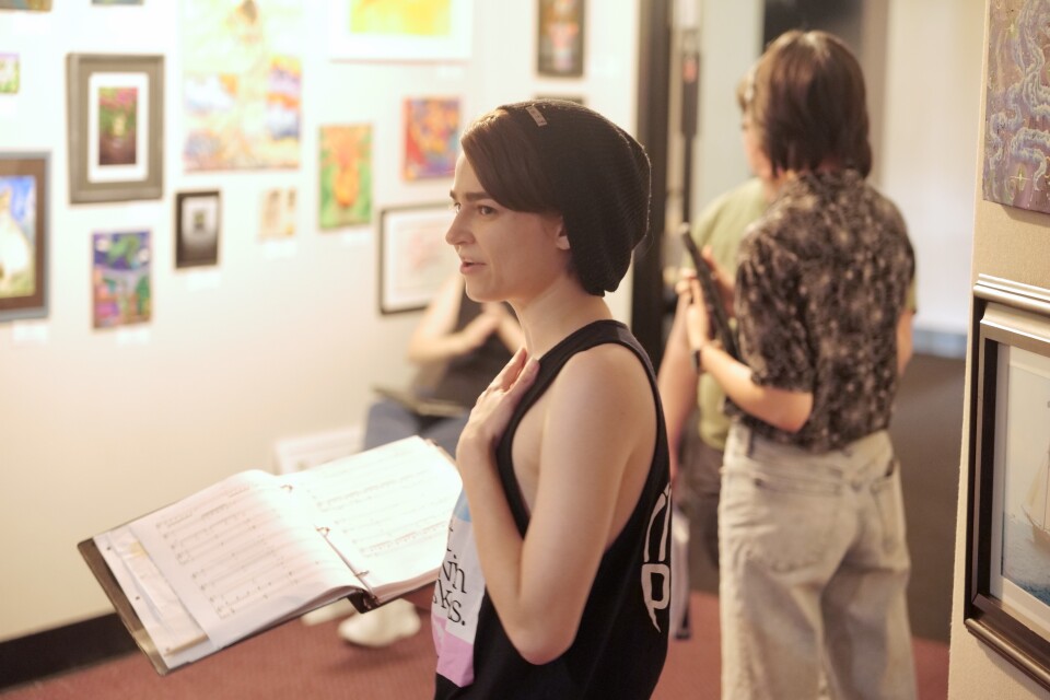 A person with short, dark hair holds a folder with sheet music. The person is in a room with art on a wall.