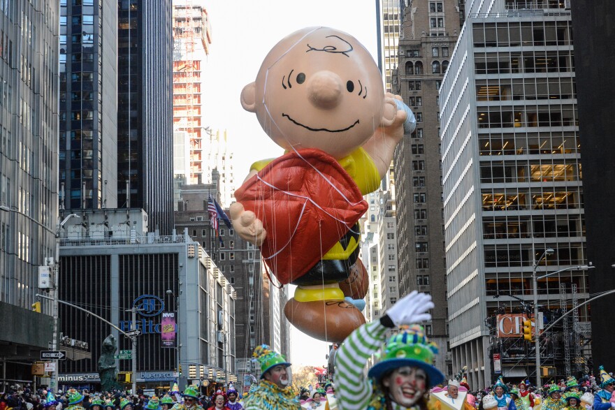 A giant Charlie Brown balloon float flies, above a downtown city crowd, connected by numerous tethers. The large float has his own giant kite in his hand, string in the other. City buildings are seen on several sides. On the ground, onlookers watch and clowns in facepaint and white and green outfits march alongside the float.