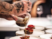 A pair of light skinned hands with several tattoos spoons a brown liquid over a series of white plates which each hold a piece of beef.
