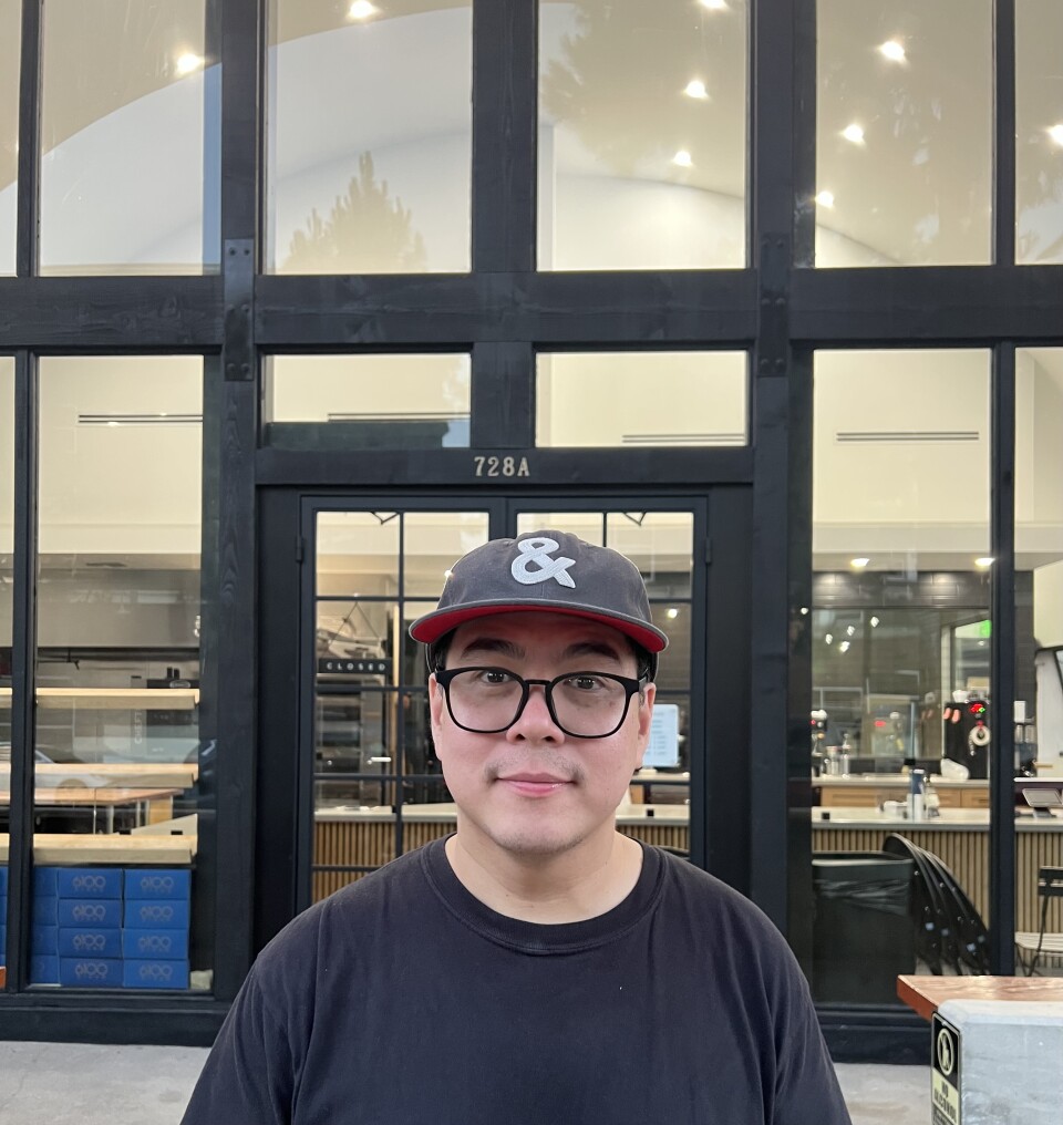 A man with medium light skin stands in front of a sleek, modern storefront with the address number 728A displayed above the entrance. The building has a minimalist design with large black-framed windows and doors, revealing a well-lit and stylish interior with clean lines, light wood, and dark metal accents.