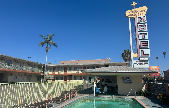 Wide shot of the Hollywood Premiere Motel showing a two-story building with mint green railings, a turquoise rectangular swimming pool in the foreground, and the iconic tall white and pink "MOTEL" blade sign with starburst design against a clear blue sky.