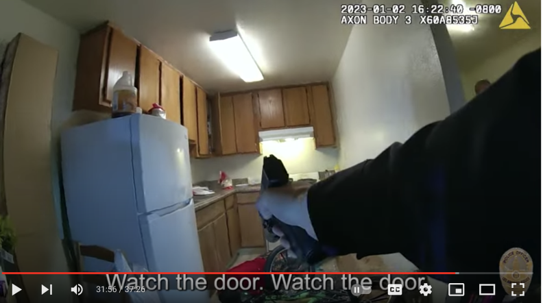 An image taken from a video that shows the arm of a police officer holding a gun, pointing the weapon into a kitchen. A refrigerator appears on one side of the room, along with light brown cabinets on the walls.