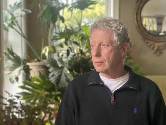 A man with light-tone skin is photographed in profile. He has gray hair and wears a pullover with a zipper.