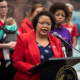 A woman wearing a red blazer and blue and white blouse stands at a podium, speaking into a microphone. A group of people stand behind her.