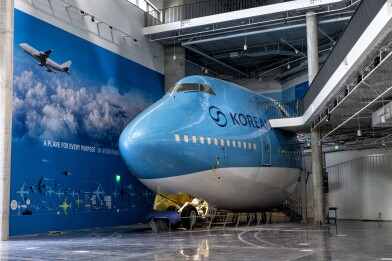 The front 70-feet of a Boeing 747 aircraft appears to be sticking out of a wall in a large room. The plane is painted mostly blue with a white underbelly and the "Korean Air" logo on the left side. A wall painted to look like the blue sky with white clouds and various small aircraft images is to the right of the plane