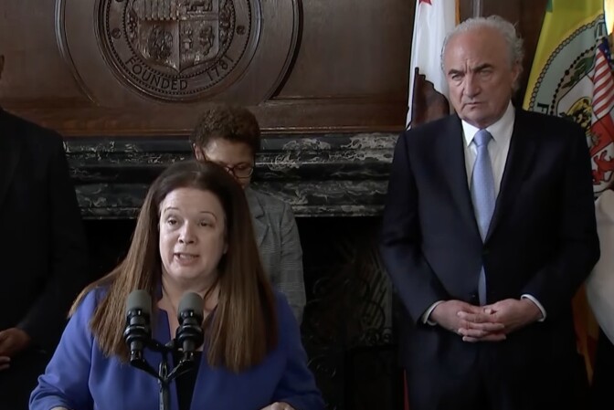 A woman with light skin tone, straight brown hair, wearing a blue blazer stands at a podium and speaks into a microphone. Behind her is Mark Adams, a man with light skin tone, white hair, wearing a navy blue blazer, white shirt, and light blue tie. He stands in front of two flags. 