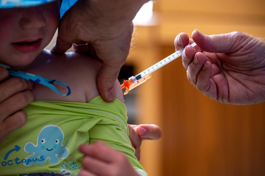 A 3-year-old boy in a green shirt and blue hat receives a COVID-19 vaccination in his left shoulder.