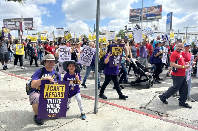 A light-skinned man wearing a tan wide-brimmed hat crouches with on a sidewalk with a sign that reads "I can't afford to live where I work." His young son also wears a wide-brimmed hat and stands to his father's left holding his hands up in a peace sign. They both wear purple shirts. Protestors march in the background. 