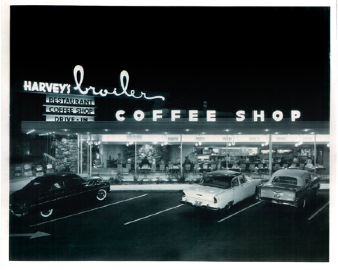 A black and white photo of cars from the 1950s parked in the parking lot of a 1950s diner.