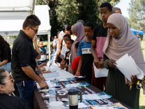A line of young people, most with dark brown skin, stand in front a display table stffed by a man wearing sunglasses and a black shirt.