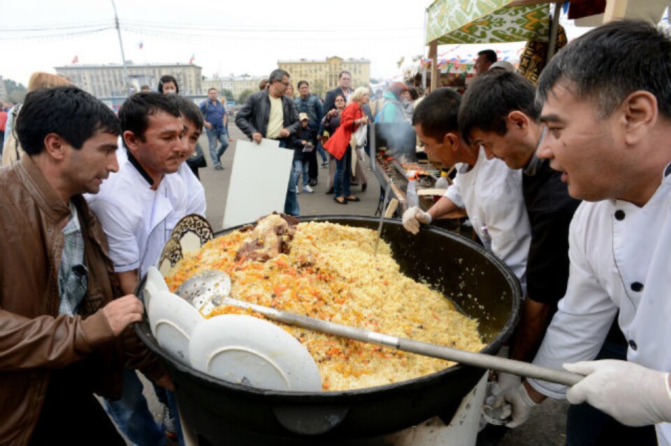 Six men sir a massive vat of rice with a long paddle. Plates are stuck into the sides of the vat.