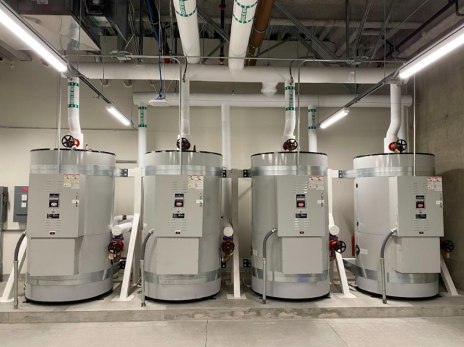 A view of four cylindrical industrial boilers inside a room with pipes coming out of them. 