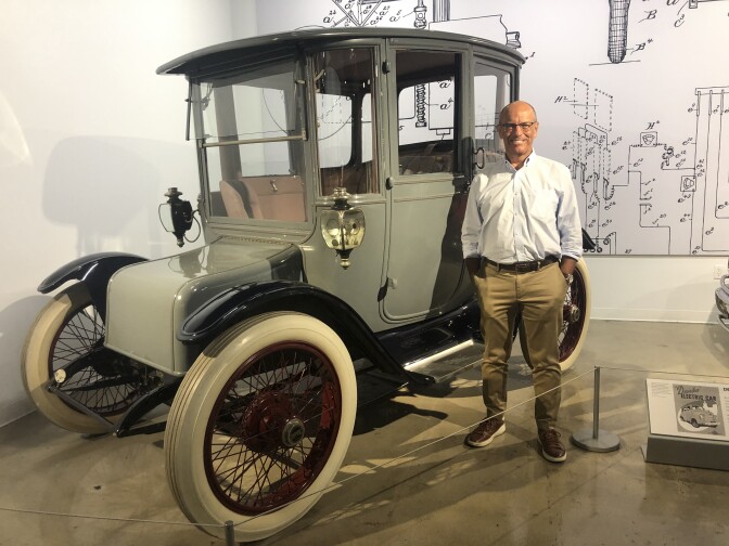 A man in a white dress shirt and khakie pants stands beside an old autombile with blue-grey finish and white spoked wheels in a museum. 