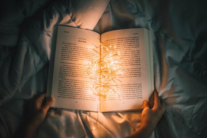A book lies open on a bed's comforter, with two hands holding each corner at the bottom. It is dark and the only light is coming from a cluster of fairy lights that lie at the crease of the open book, illuminating the words. 