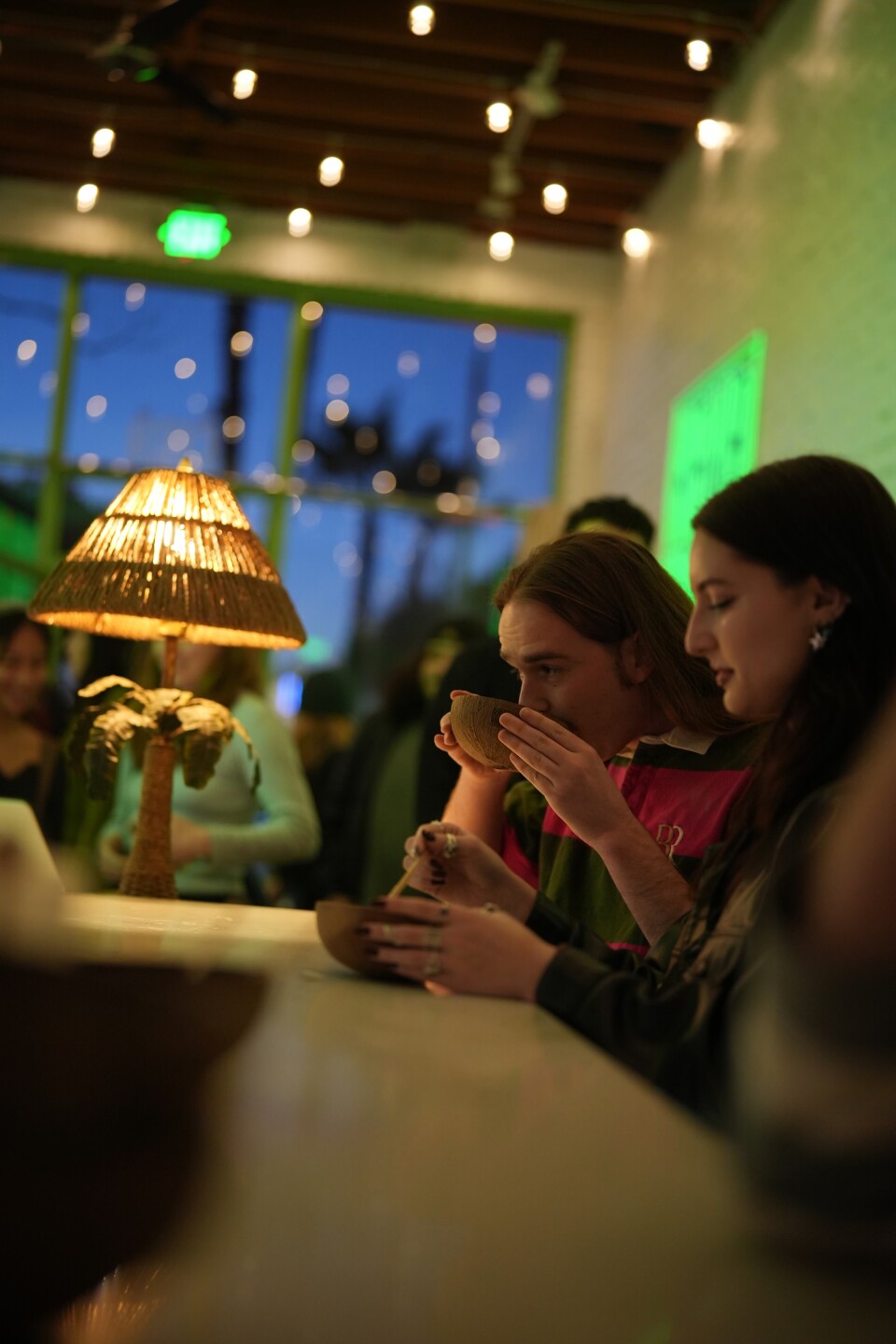 Two light-skinned individuals are seated next to each other at a bar with dim lighting. One is sipping from a cup made out of half a coconut shell. The other is holding an identical cup on the bar. In the background is a light with a woven lampshade and a palm tree serving as its base.