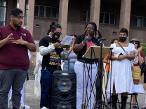 Amber Sam stands at a podium surrounded by a crowd of people who attended a rally to call on LA County to invest more in mental health treatment beds. Sam wears white pants and a black shirt. 