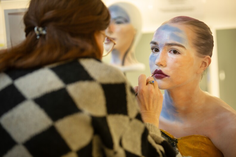 A young woman is getting makeup applied to her face and lips. Her forehead and neck are blue and white, her lips are deep red.