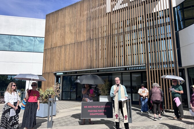 A man wearing a cream-colored stole speaks into a microphone in front of a building. A group of people stand in a semi-circle around him, several holding umbrellas to shield them from the sun. 