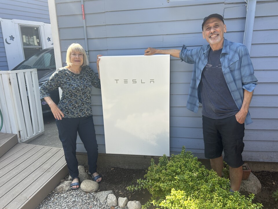 An older white woman with a white bob, blue flower pattered shirt and blue pants stands to the left of a large white rectangular Tesla battery on the outside wall of their blue-painted buildig. On the right, am older white man wears a blue flannel shirt over a blue t-shirt and black shorts. They both smile. 