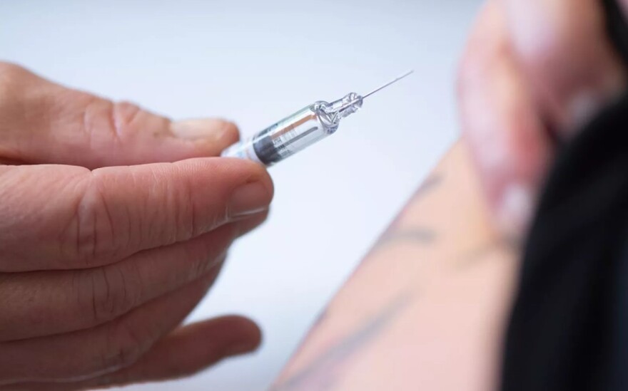 A hand holds a syringe as a blurry image of an arm in the background pulls up a shirt sleeve.