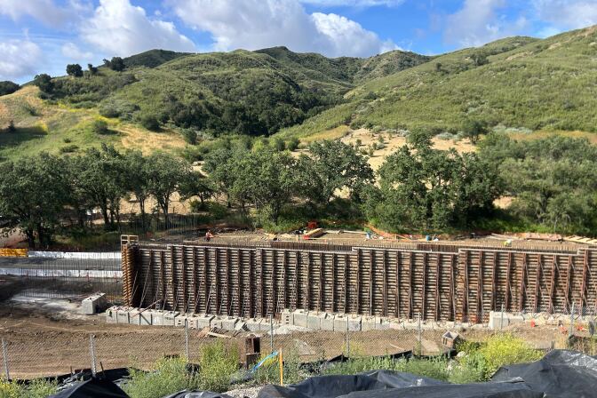A wall with scaffolding stands in front of a hillside lined with trees and grass.  