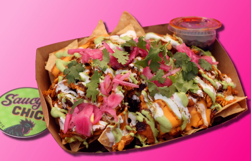 set against a bright pink background, a brown cardboard food container holds a pile of tortilla chips, meat, pickled onions and white sauce.