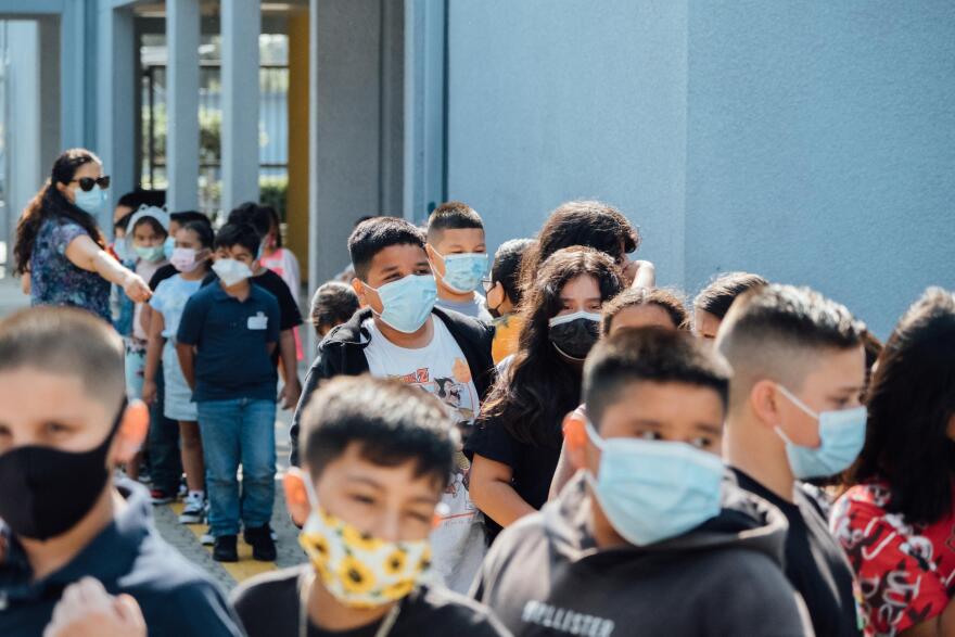 Kids stand lined-up in a school yard wearing masks.