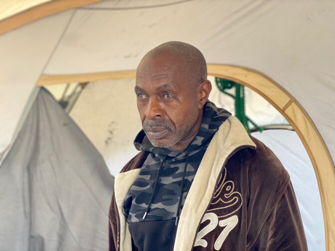 Banyard is shown in front of his tent looking off camera. He is wearing a camouflage hoodie and a brown jacket.