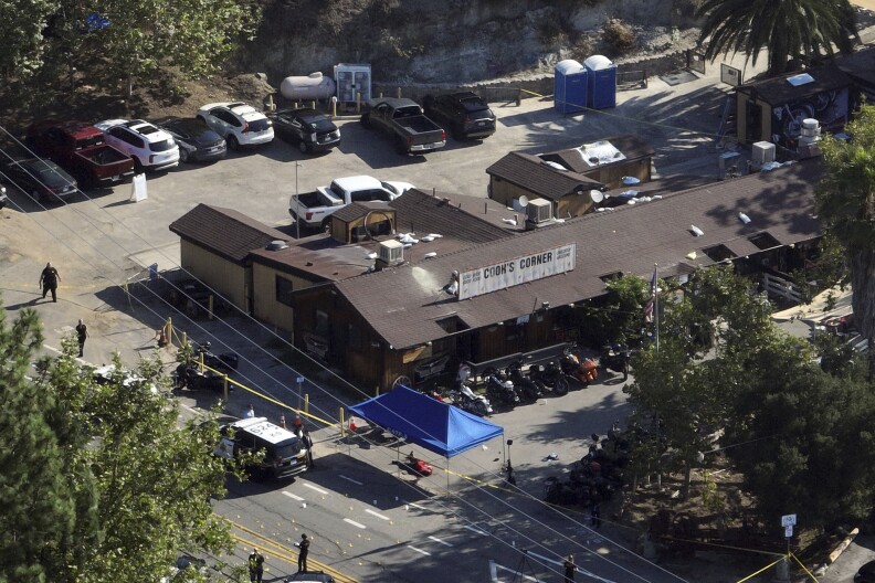 A bird's eye view looking down at a large one-story building with sloping wood-shingled roof. A banner-like sign on the roof reads "Cook's Corner." A police squad car is parked out front near a blue tarp and along one wall of the building is a row of motorcycles.