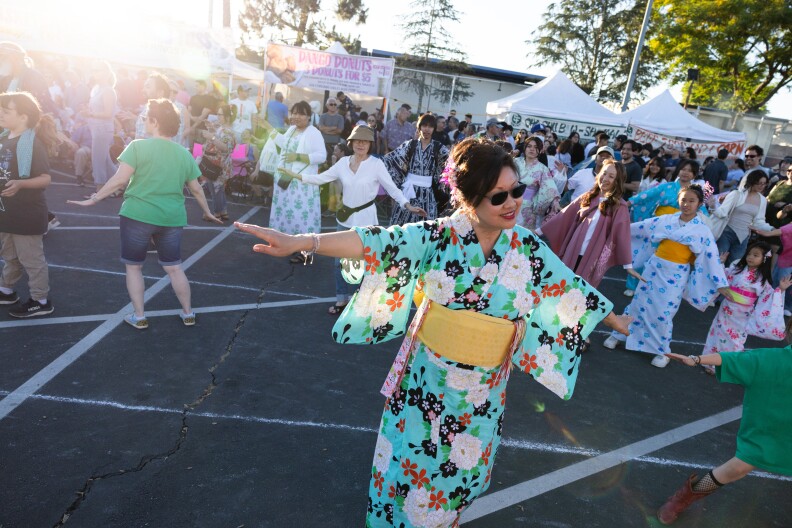 An Asian American woman wearing a teal yukata with her hair up and sunglasses, spreads out her arms. Behind her, a line of people copy her movements.