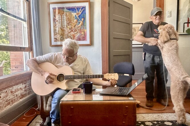 An elderly man with white hair plays guitar, while a large goldendoodle dog jumps on a hispanic older man wearing a hat. 