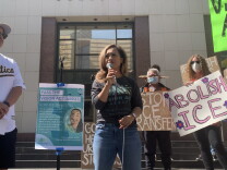 An Asian American woman stands on the steps of a building in downtown holding a mic as people around her hold signs that read "Abolish ICE" and "Stop ICE Transfers."
