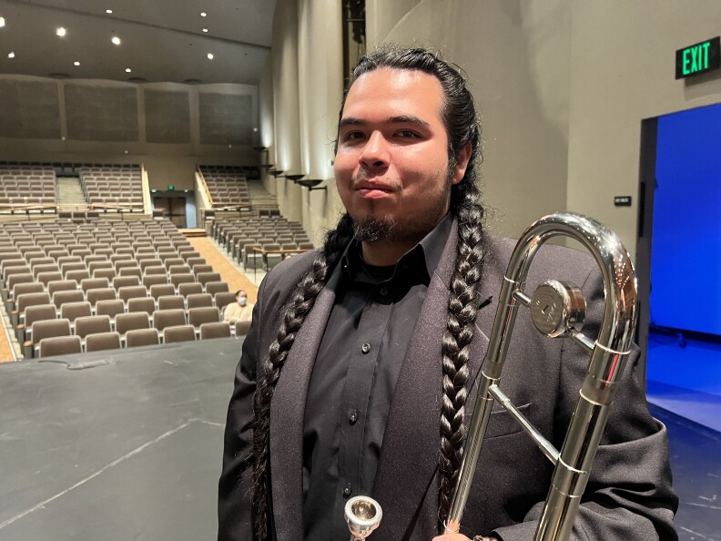 A young man with light brown skin and long braids stands with his tuba on stage.