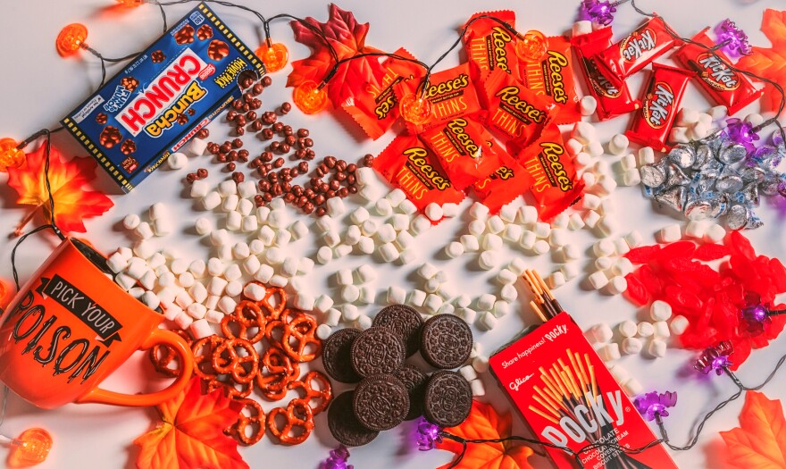 An array of candy is spread out on a table decorated with Halloween decor. Candy includes Crunch, Pocky, Reese's, KitKats and Oreos, as well as pretzels and marshmallows.